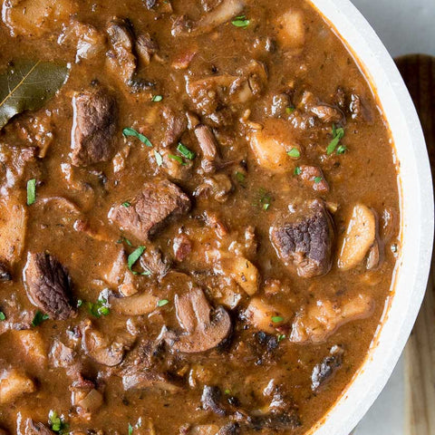 A bowl of Beef & Mushroom Stew with visible chunks of beef and mushrooms in a brown gravy-based sauce, garnished with chopped parsley.