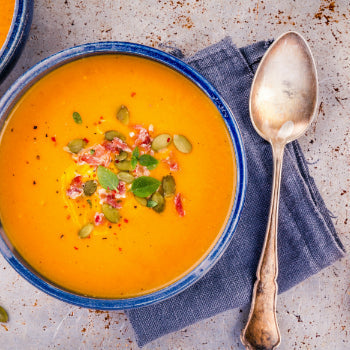 A bowl of butternut squash soup garnished with seeds and herbs, with a spoon on the side and ingredients visible in the background.