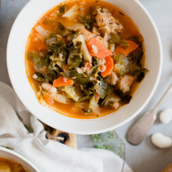 A bowl of Smoked Chicken Sausage, White Bean & Kale Soup with visible ingredients including carrots, kale, and white beans.