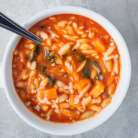 A bowl of fire roasted tomato and vegetable soup with orzo and romano cheese, with a spoon partially visible in the bowl.