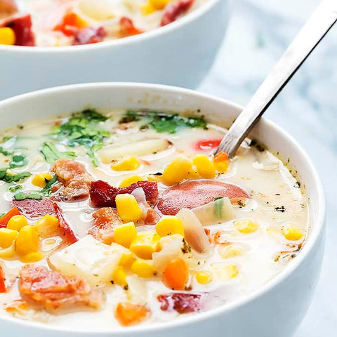 A bowl of Turkey Bacon & Vegetable Chowder with a spoon, featuring ingredients such as potato, carrot, corn, and zucchini, garnished with parsley.