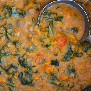 A ladle full of West African Sweet Potato & Peanut Stew, showing a thick, creamy soup with pieces of sweet potato, carrot, and spinach.